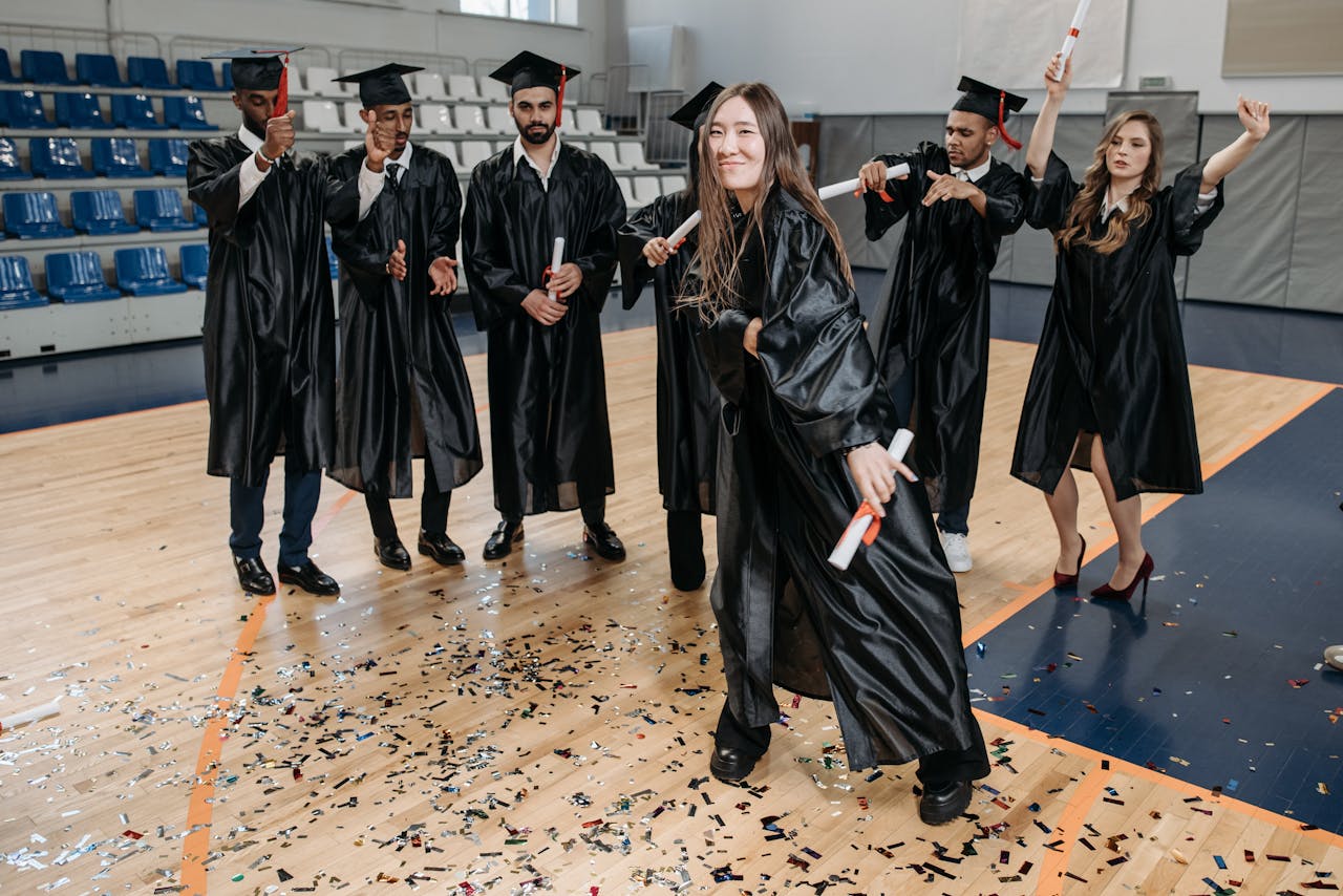 Happy graduates celebrating indoors in a gymnasium with diplomas and confetti.